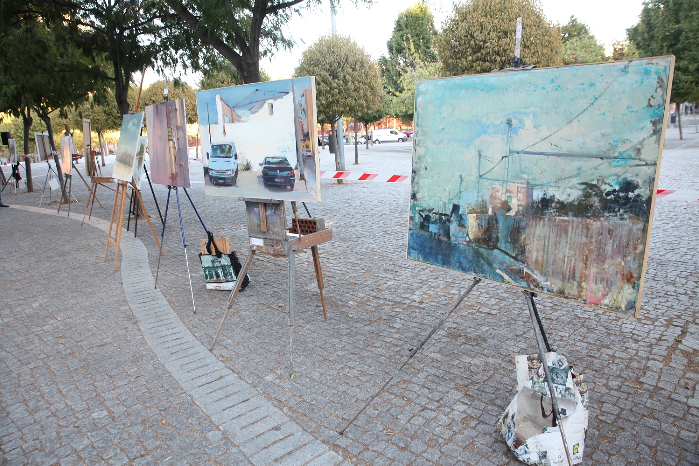 Media docena de  lienzos expuesto en una plaza con árboles 