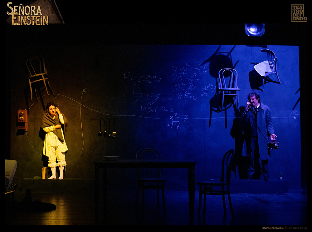 Un hombre y una mujer en un escenario de teatro, oscuro y con sillas y teléfono