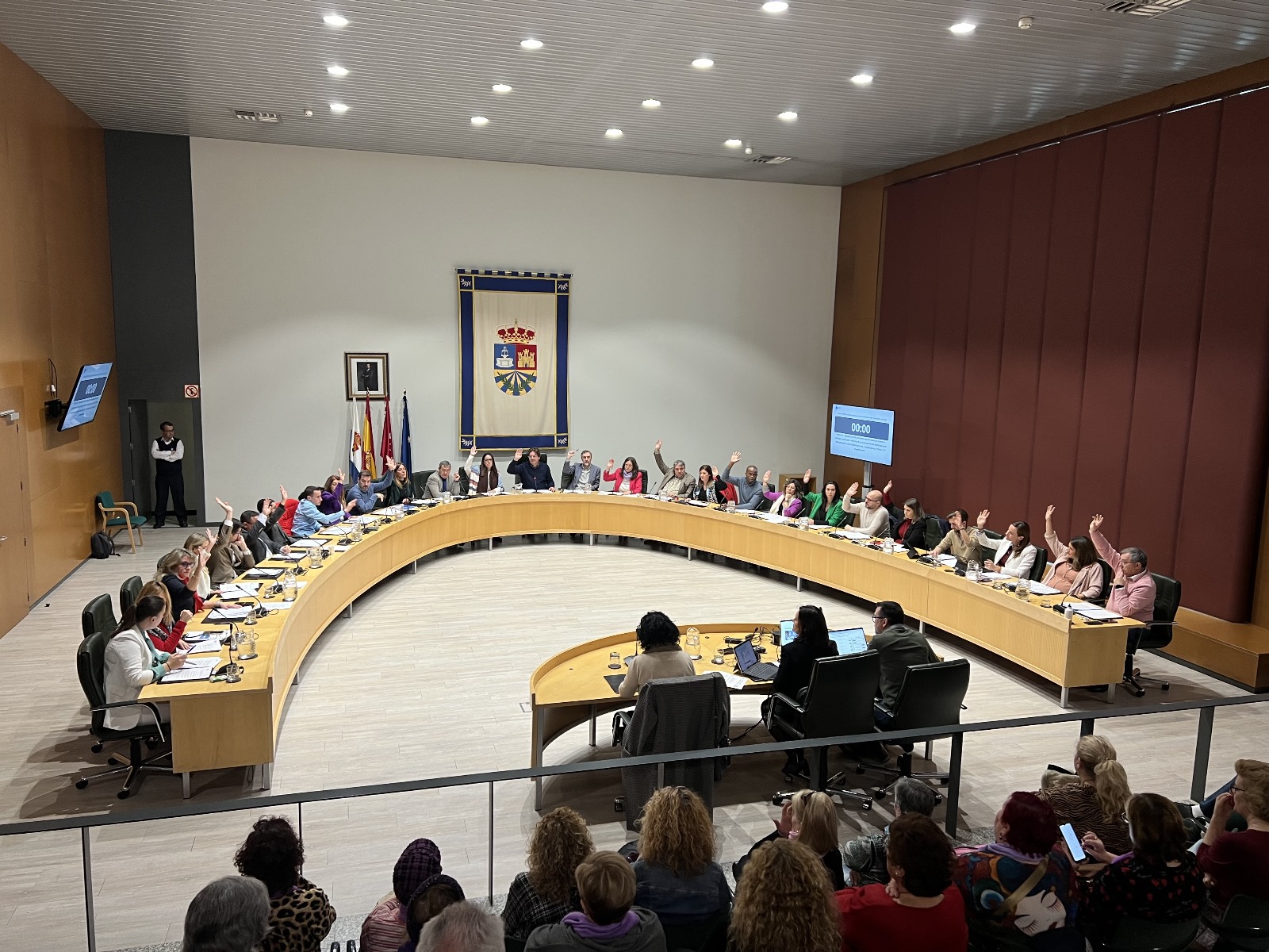Una veintena de hombres y mujeres sentados en torno a una mesa en semicírculo con el brazo en alto