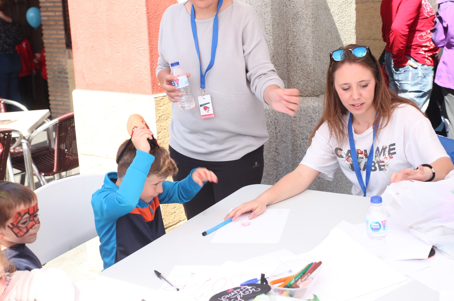 Un niño pequeño pintado la cara de rojo y negro y otro quitándose una careta sentados a una mesa con dos mujeres haciendo juntos manualidades