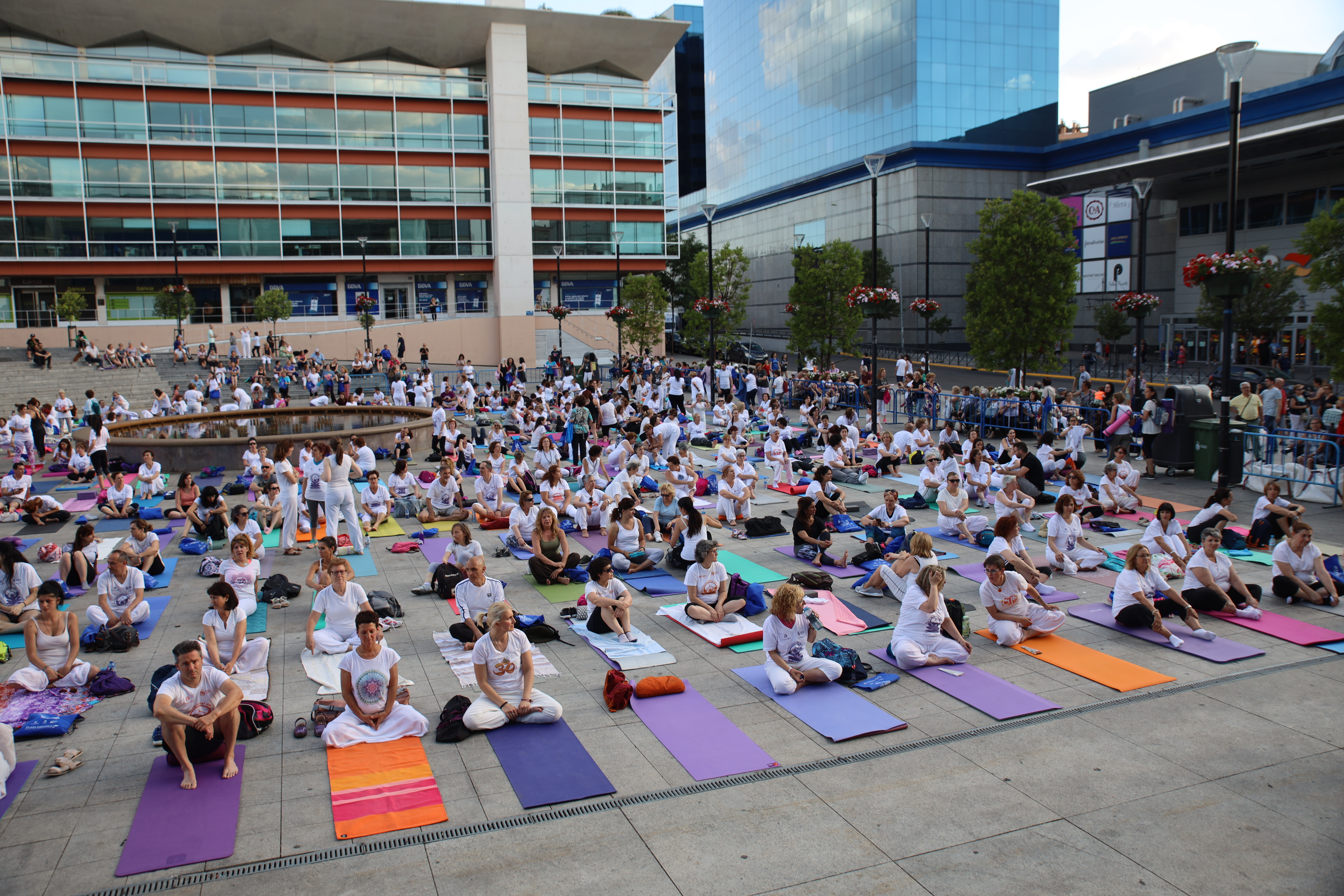 Un centenar de personas sentadas en esterillas se preparan para una clase de yoga en una plaza al aire libre
