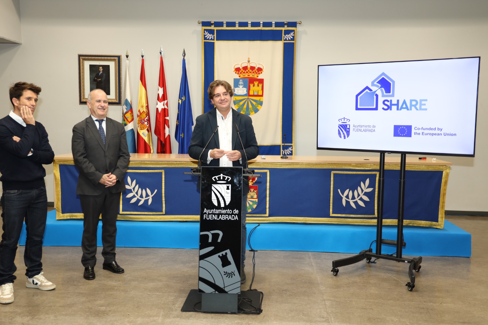 Tres hombres , uno de ellos habla por un micrófono. Al fondo banderas, un tapiz con escudos una televisión