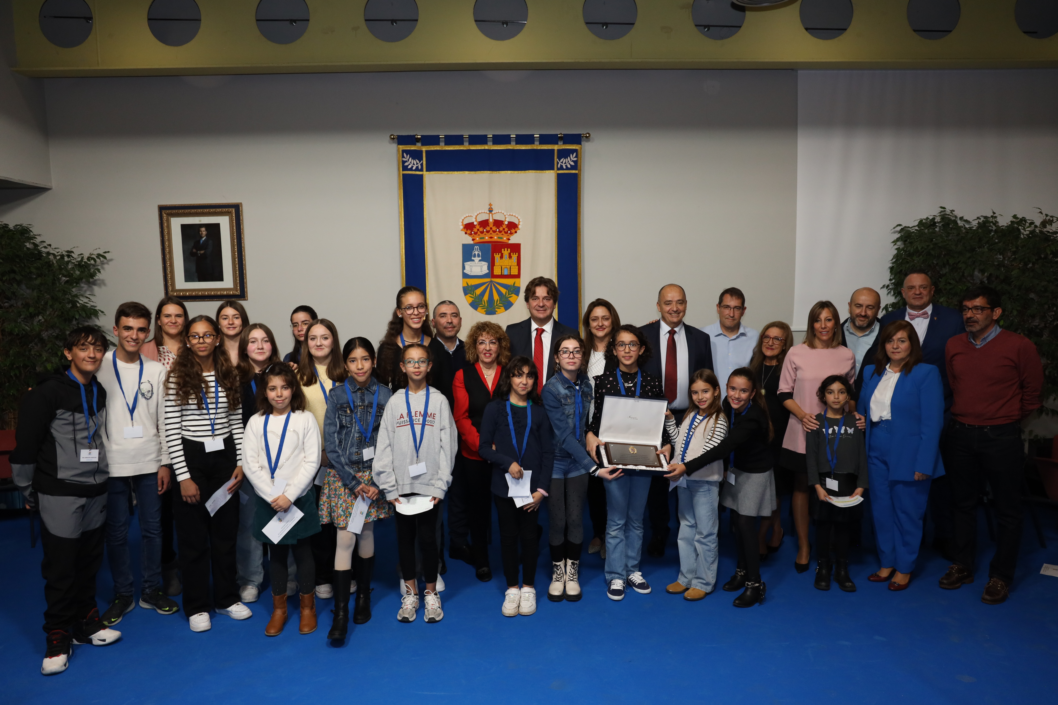 Una veintena de niños, niñas y personas mayores delante de un escudo de tela 