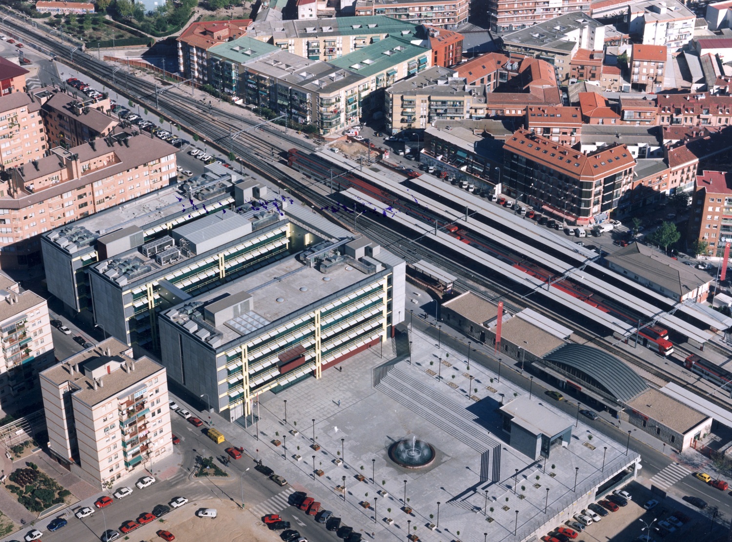 Vista aérea de una plaza cuadrada y grande con suelo gris y a no de sus lados tres edificios, al fondo vías de tren 